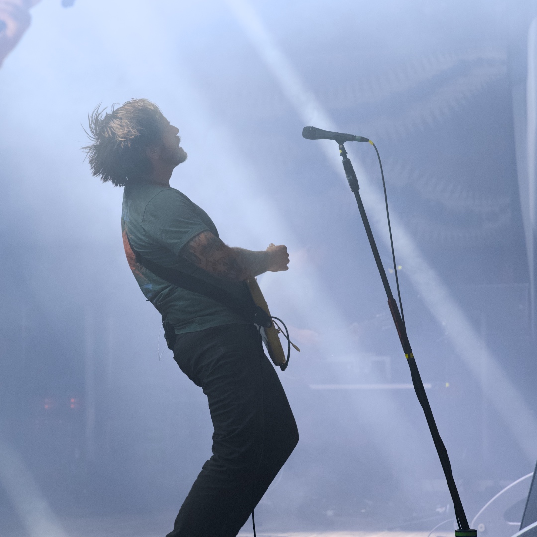 Dustin Kensrue, the guitar player from Thrice, playing his guitar with a microphone stand in front of him and bright lights behind him.
